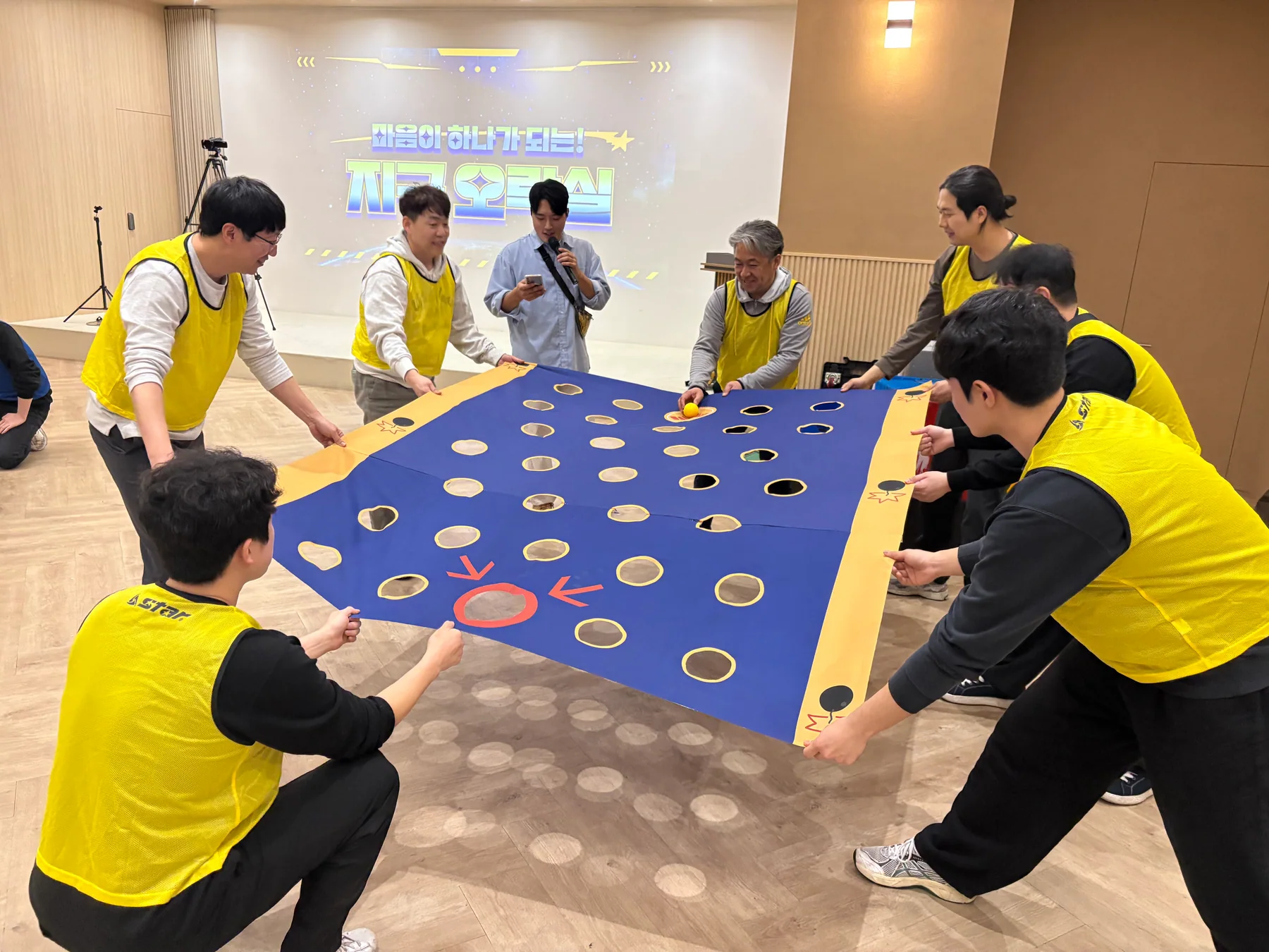 노란 팀 참가자들이 구멍이 뚫린 대형 천을 함께 잡고 공을 골대까지 이동하는 실제 팀빌딩 현장